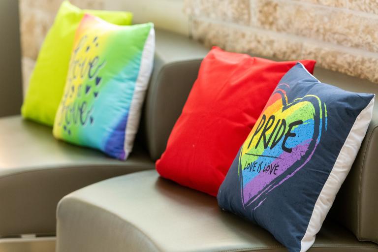 Four colourful cushions on a bench with Pride written on the first one. Partially obscured.