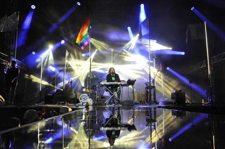 A performer is sitting behind a keyboard on stage with beams of blue and white lights and a rainbow flag flying on a pole. Partially obscured.