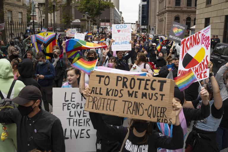 A group of people crowd into a city street holding signs and rainbow flags. The sign in the foreground reads, “To protect all kids we must protect trans and queer kids.”