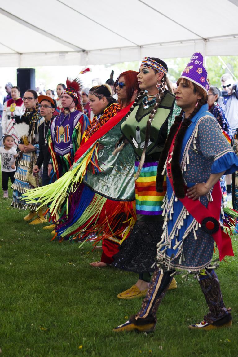 A group of people wearing colourful Indigenous regalia dancing in a line inside a tent on grass.