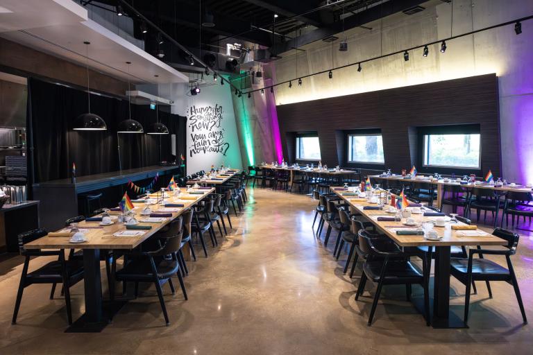 An open concept restaurant features rows of rectangular wood tables covered with plates and rainbow flags. Partially obscured.