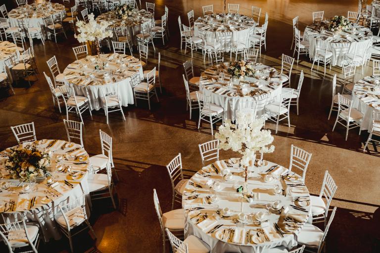 Round dinner table with white linen, white chiavari chairs and white flowers on it, set with cutlery. Partially obscured.