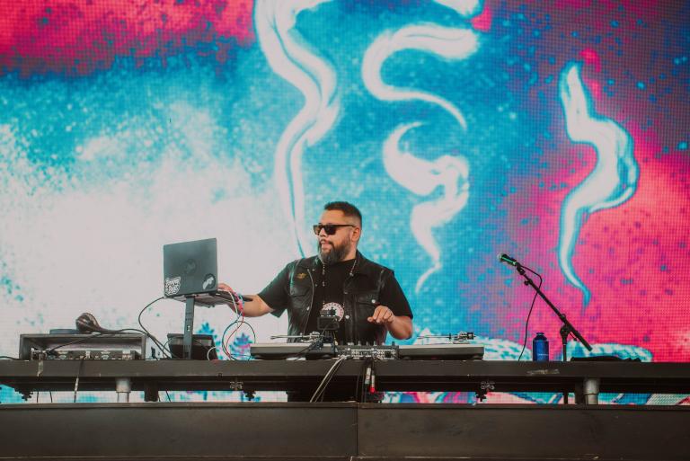 A bearded, dark-haired man, wearing sunglasses and a black t-shirt, is performing on stage using a computer and an electronic keyboard mixer. Partially obscured.