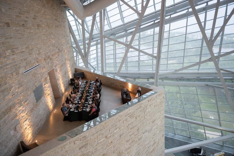 An elegant long table with people dining in an architecturally striking space, featuring tall glass windows and a high ceiling supported by metal beams.