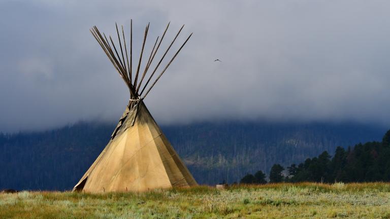 A large tipi sits in a field of grass. Its poles rise into a misty sky. Partially obscured.