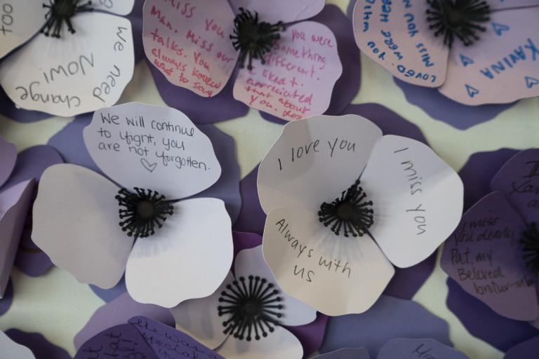 Three-petal flowers made of paper in various shades of purple are grouped together in a wall display. Names and messages are written on them. Partially obscured.