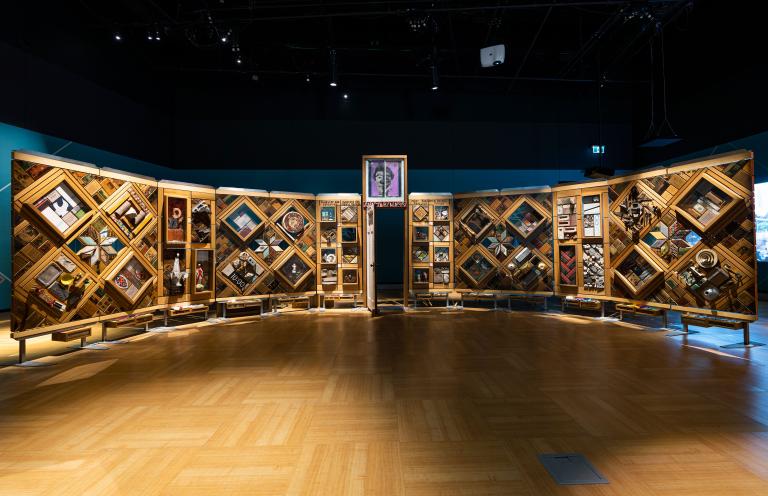 A large, curved freestanding wooden wall on which is mounted numerous small objects arranged in diamond and rectangular patterns. There is an open door in the centre of the wall. Partially obscured.