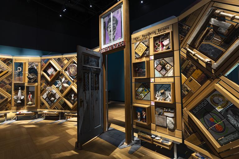 A large, curved freestanding wooden wall on which is mounted numerous small objects arranged in diamond and rectangular patterns. There is an open door in the centre of the wall. Partially obscured.