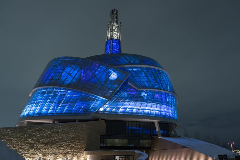 The exterior of the Canadian Museum for Human Rights is pictured at night. The building is lit up from the inside with vibrant deep blue lights. Partially obscured.