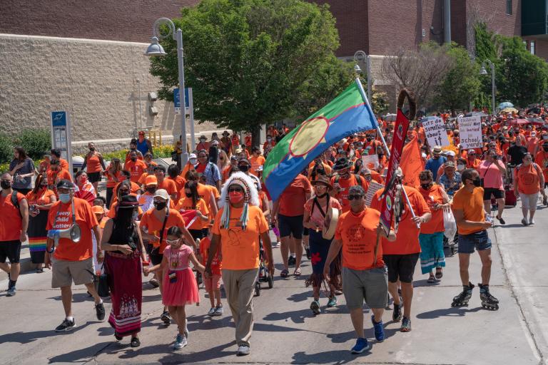 Hundreds of demonstrators in orange shirts, many also wearing traditional Indigenous articles such as a headdress or ribbon skirt and carrying signs, march down a wide paved street. Partially obscured.