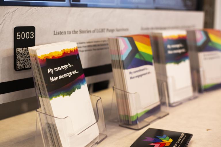 Display of cards with rainbow designs and messages, set in clear holders on a table. A poster in the background reads, “Listen to the Stories of LGBT Purge Survivors.”
