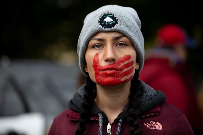 A young woman with braided hair, wearing a maroon jacket and gray winter cap, looks forward resolutely. A red handprint is painted over her mouth and face. Partially obscured.