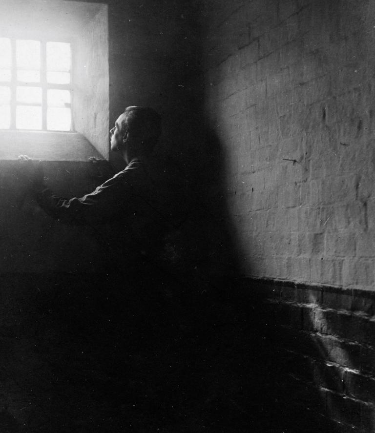 A man stands in dark shadows in a stark stone cell, gazing upward at bright sunlight streaming in through a barred window. Partially obscured.