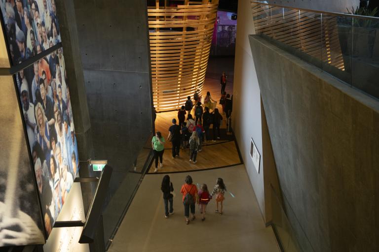 Students walking through Museum galleries. On the left wall is an image of a crowd of people. The walls are made of cement. Partially obscured.