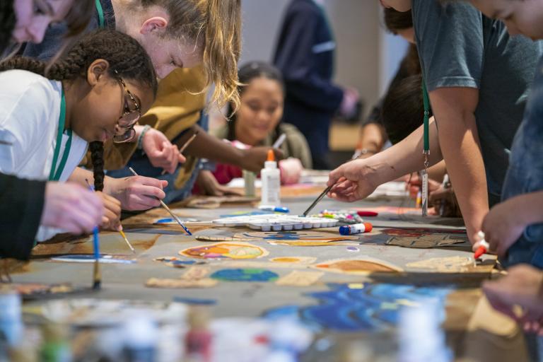 A group of children and youth paint with small brushes. They are gathered on both sides of a long table with different coloured paints in the foreground. Partially obscured.
