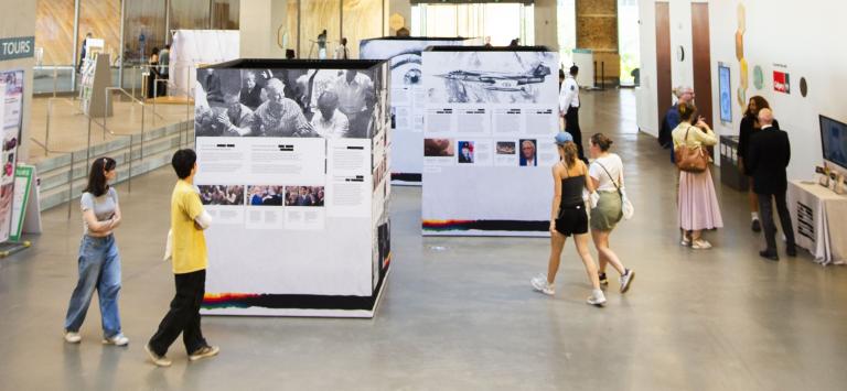 Three large cubes covered in images and text are on display in a large public room. Two groups of two adults walk near them, perusing the cubes’ content. Off to the side, four people are talking.