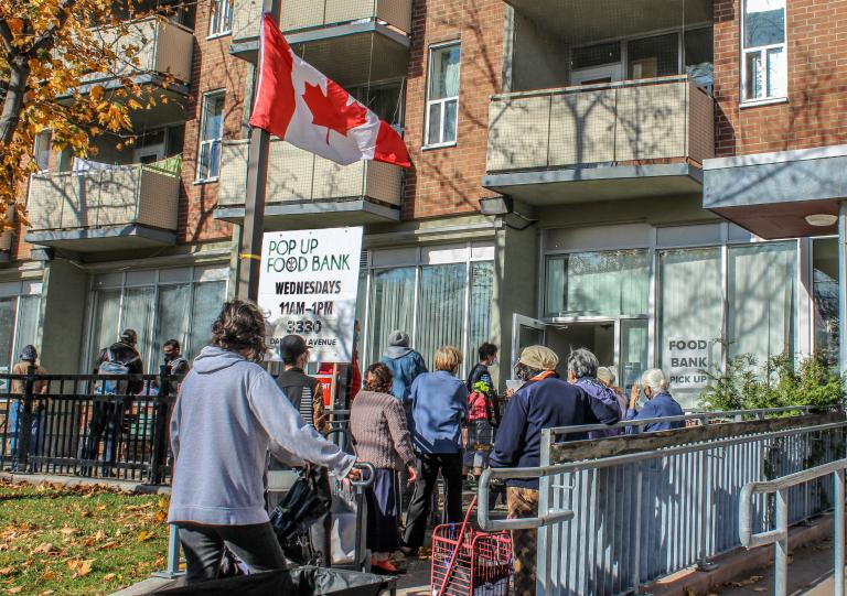 A line-up of people wait outside a building. There are signs indicating it is a Food Bank. A Canadian flag flies above them. Partially obscured.