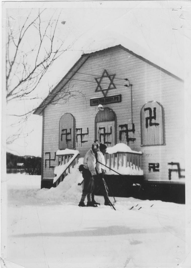 Two people play hockey in front of a synagogue with several swastikas painted on it. Partially obscured.