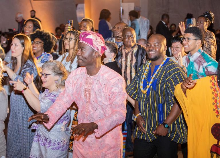 A group of people of all ages, many in brightly coloured traditional African attire, dancing and smiling. Partially obscured.
