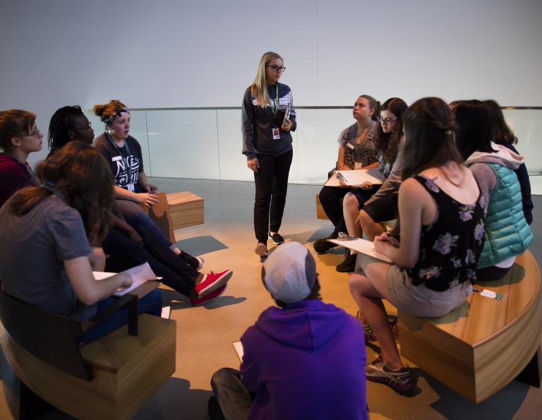 Students sit on benches in a circle with clipboards on their laps. A woman is standing and talking to them. Partially obscured.