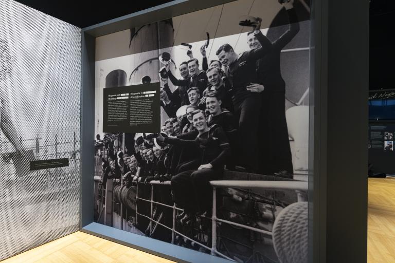 In a museum display case, a large black and white photo shows a group of navy personnel smiling for the camera. Partially obscured.