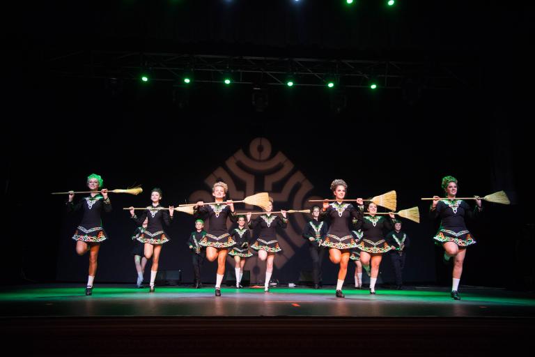 Thirteen Irish dancers dressed in short, black dresses are holding brooms across their chest. Partially obscured.
