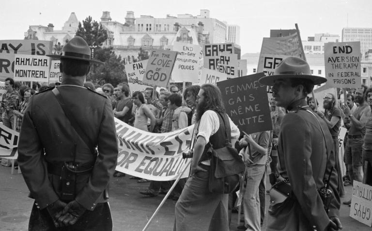 A large group of protestors march through a city while two uniformed RCMP officers keep watch. In this black-and-white image, the protestors' signs and banners call for an end to discriminatory practices against members of the 2SLGBTQI+ community. Partially obscured.