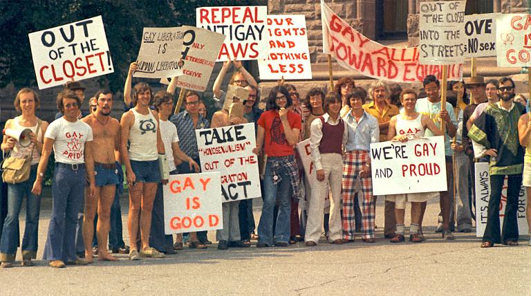 A large group of people dressed in T-shirts and seventies-style flared pants, stand together holding signs with messages such as “Out of the Closet,” “Gay is Good” and “Repeal Anti-Gay Laws.” Partially obscured.