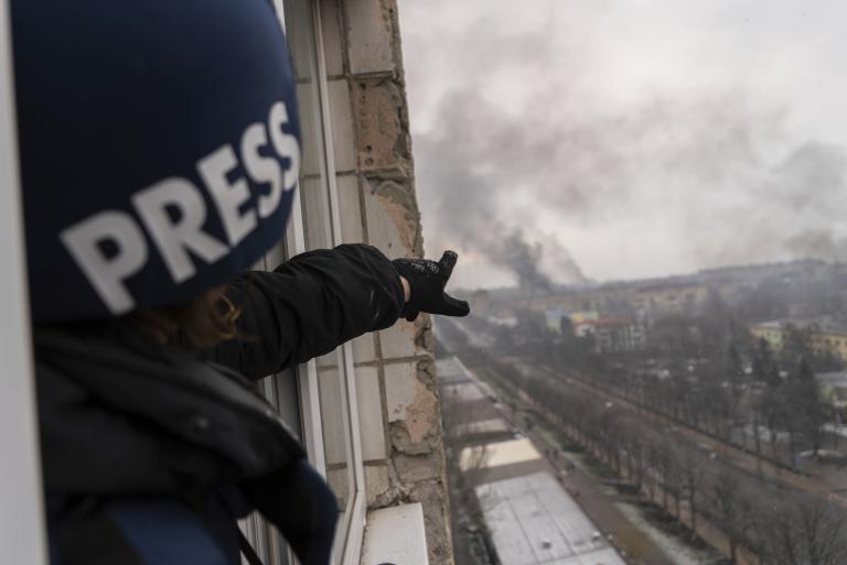 A person wearing a dark blue helmet marked with the word “PRESS” points from a balcony window high above a road, towards a pillar of smoke following an explosion. Partially obscured.