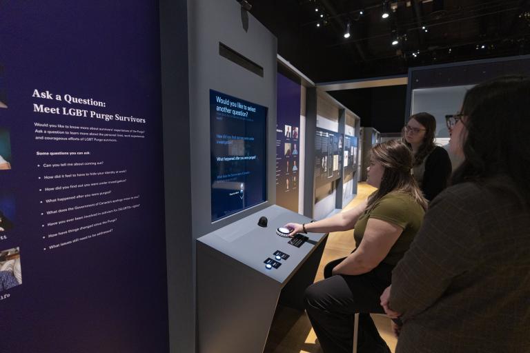 Three people gathered around the “Ask a Question: Meet LGBT Purge Survivors” video interactive station. Partially obscured.