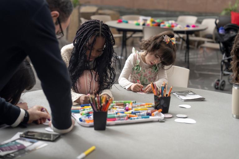 Two children sit at a table making art while an adult leans on the table, watching. Partially obscured.