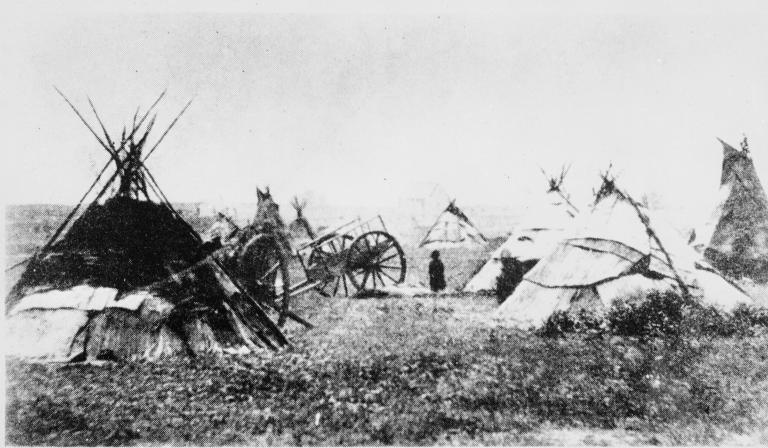 A grainy black and white photo of several tipis and a wooden cart in a field. Partially obscured.