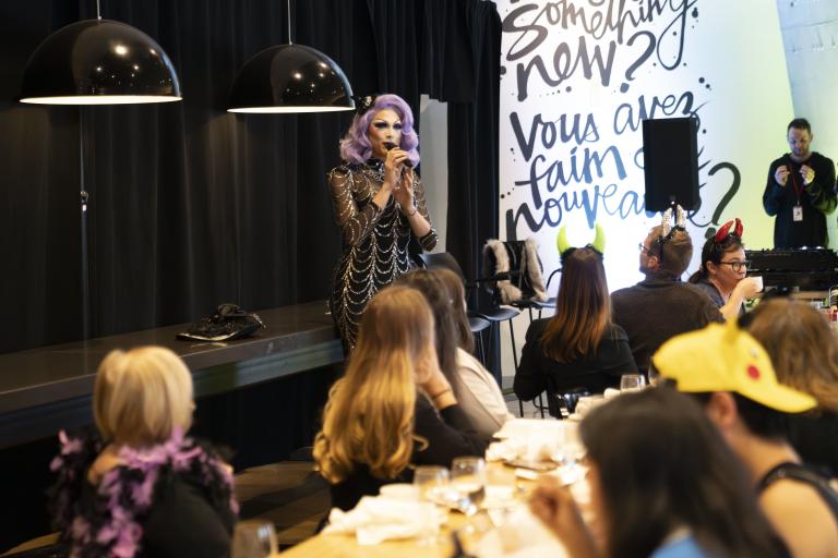 A drag performer with purple hair and a silver and black dress speaks in front of a group having brunch. Partially obscured.