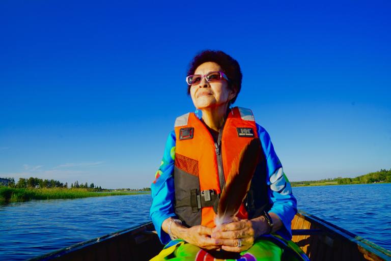 An indigenous woman is in a canoe on a lake with an orange life jacket. She seems at serene. Partially obscured.
