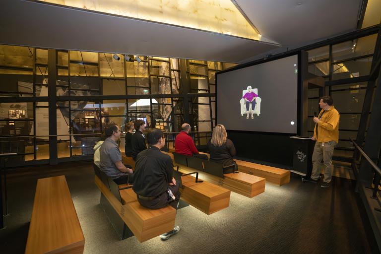 A small group of people sitting on wooden benches in a theatre face a screen showing an older woman seated in a chair. A man in a yellow shirt is standing to the right of the screen holding a microphone. Partially obscured.