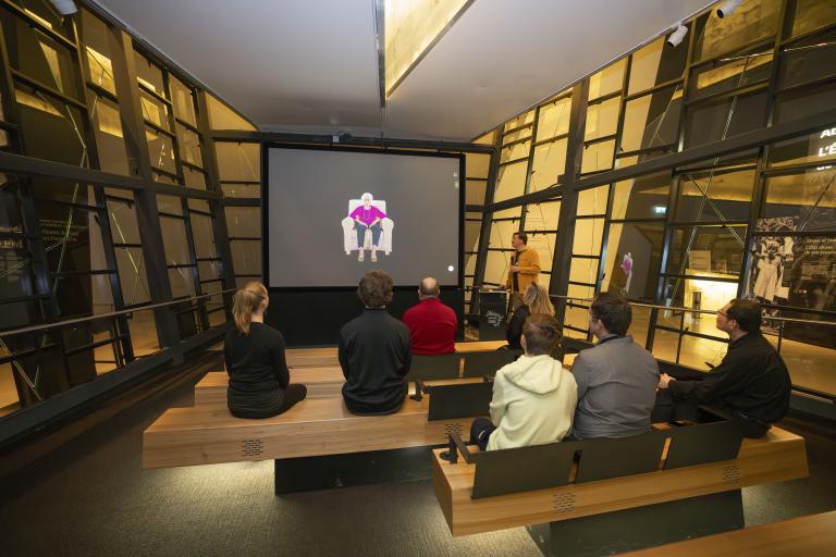 A small group of people sitting on wooden benches in a theatre face a screen showing an older woman seated in a chair. A man in a yellow shirt is standing to the right of the screen holding a microphone. Partially obscured.