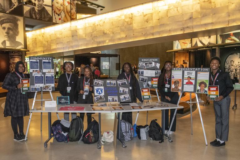 Six black students in a museum hall present proudly their work on posters and on a table. Partially obscured.