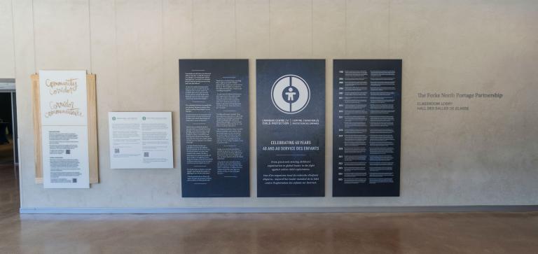 Large dark blue panels with white text and graphics displayed in the Community Corridor at the Canadian Museum for Human Rights. The centre panel features a circular logo and commemorates the 40th anniversary of the Canadian Centre for Child Protection. The two panels on either side present bilingual text in English and French. Partially obscured.