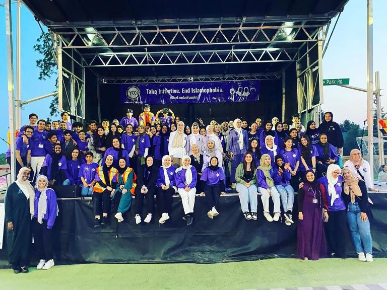 Dozens of people are gathered on a stage, most smiling and wearing the colour purple. Many are wearing hijabs. A banner hangs at the back of the stage that reads "Take Initiative. End Islamophobia. #OurLondonFamily." Partially obscured.
