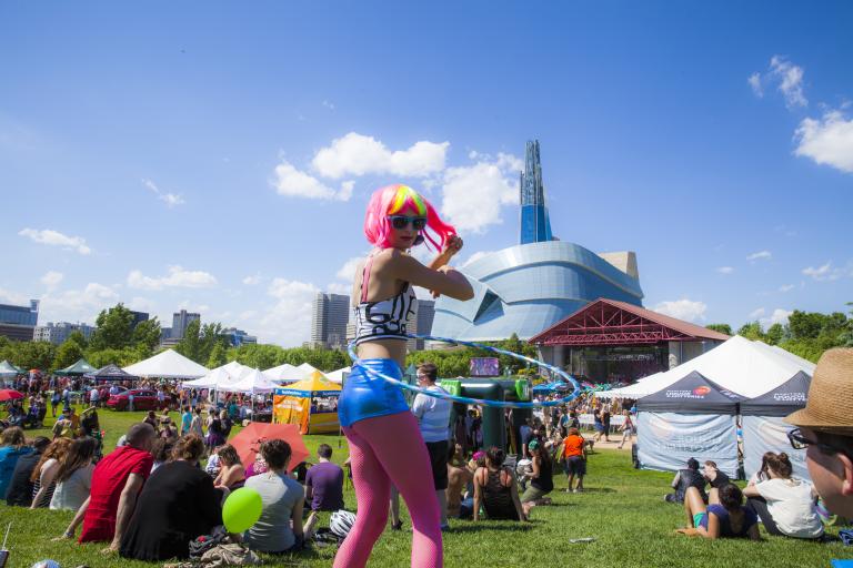 A hula hoop dancer with a pink and fluorescent yellow wig with pink leggings walks in a crowd of people sitting on a field of grass in front of a large glass building. Partially obscured.