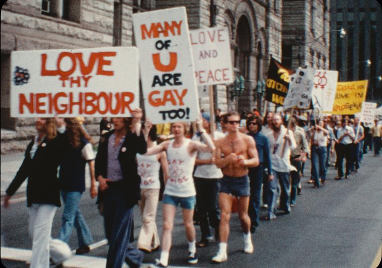 A group of people march down a city street. At the front of the group, on the left side of the image, two people hold a sign over their heads that reads “Love thy neighbour.” Slightly behind them is a sign that reads “Many of u are gay too!” Partially obscured.