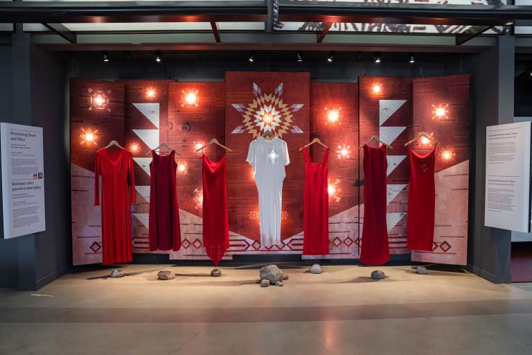 Six red dresses and a white dress in the middle are suspended on wooden clothes hangers in front of a backdrop. A white star glows at the location of the heart on the central white dress. The backdrop features patterns on a birchbark texture. Some of the patterns are illuminated from behind to create glowing star-like features on the panels. Partially obscured.