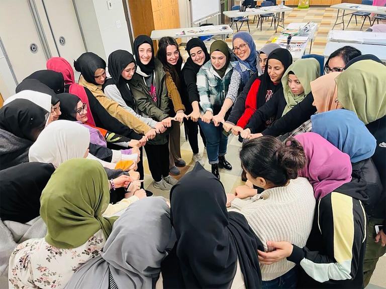 A group of smiling Muslim women in a circle with linked hands. Partially obscured.