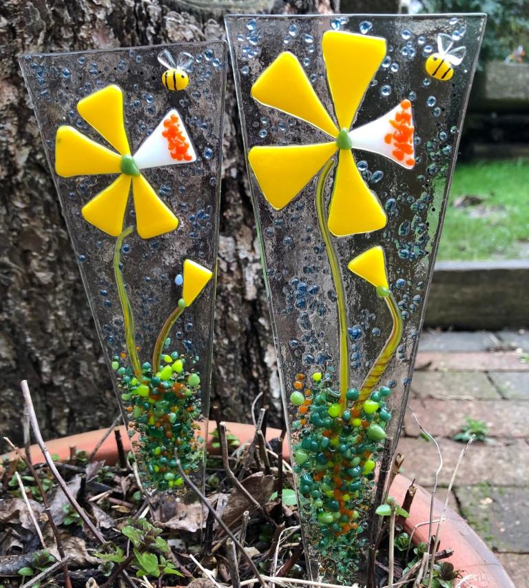 Two fused glass ornaments depicting yellow flowers sit in a flower pot outside. Partially obscured.