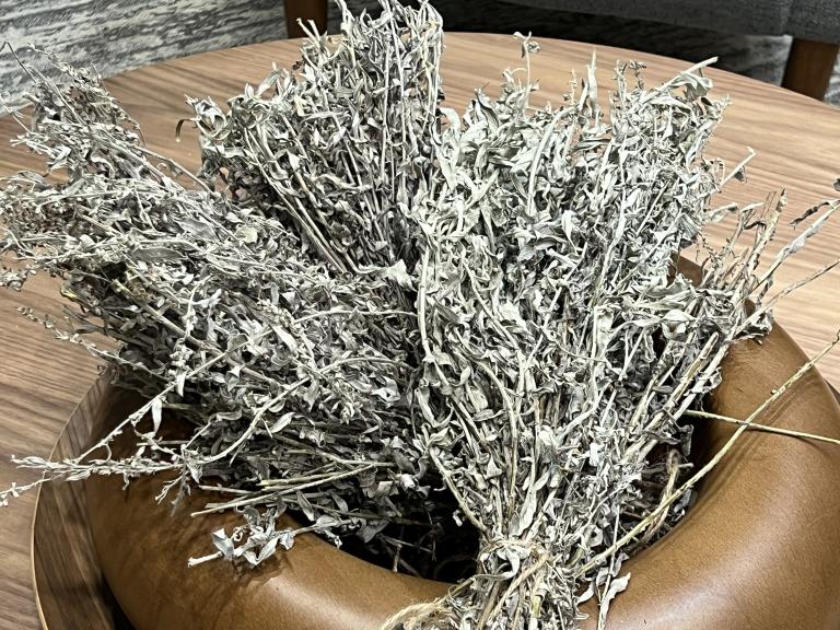 A bundle of sage, tied at the stalk, sits in a small wooden bowl. Partially obscured.