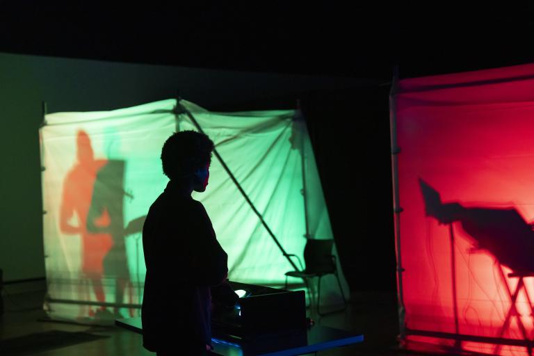 The silhouette of a person standing in the dark in front of red and green clothed box structures lit from the inside and containing shadowed figures. Partially obscured.