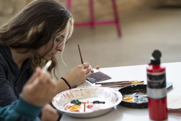 A young woman sitting at a table paints a design on a tile while a tin tray with various paints sits in front of her. An open bottle of red paint sits on the table. Partially obscured.