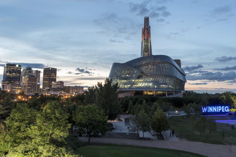 Photo extérieure des fenêtres en verre et de la tour du Musée canadien pour les droits de la personne, prise au crépuscule avec le ciel des prairies en arrière-plan. Le panneau Winnipeg à Forks est situé à droite de l'image. Plusieurs tours de bureaux et les lumières du centre-ville sont visibles à gauche de l'image. Partially obscured.