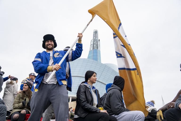 A smiling man in a crowd of people holding a large yellow flag with a blue and white, “W” in the centre. The man is wearing a blue Winnipeg Blue Bombers jacket as he stands in front of the Canadian Museum for Human Rights. Partially obscured.