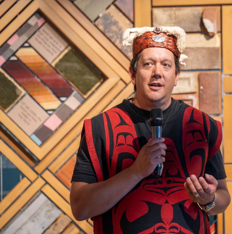 A man stands with a mic speaking to a crowd wearing traditional indigenous clothing infront of a wood panel art installation. Partially obscured.